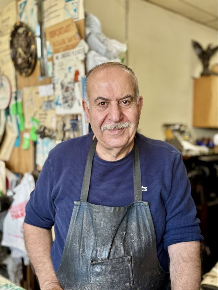 George at the workbench of his shoe repair shop, smiling in a navy shirt and leather work apron